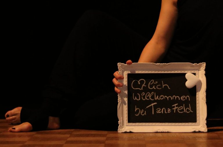 Schwarze Tafel mit weißer Schrift, gehalten von einer Person in Schwarz, im Hintergrund dunkel.