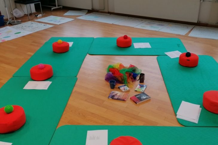 Orangefarbene Sitzkissen in einem Kreis um Bücher und Materialien auf dem Boden.