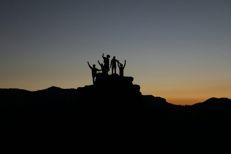 Silhouetten von Personen auf einer Bergspitze. Im Hintergrund der Sonnenuntergangshorizont.