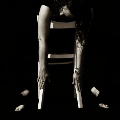 Schwarz-weiß Aufnahme von einer Frau mit langen Jahren, die sich falsch herum auf einem Stuhl sitzend den Kopf auf die Lehne liegt.