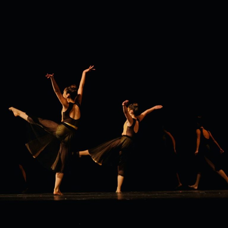 Tanzende Frauen in schwarzer Kleidung vollführen dynamische Bewegungen auf dunkler Bühne. 