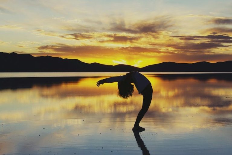 Person in einer Yoga-Positur vor einem Sonnenuntergang in einem See stehend.