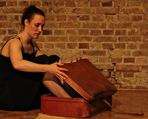 Frau in schwarzem Kleid sitzt vor einer Ziegelwand und öffnet einen Koffer.