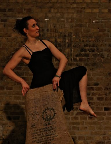 Tanzende Frau in schwarzem Kleid, steht mit einem Bein in einem alten großen Kaffeesack.